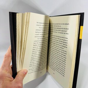 May include: An open book with cream-coloured pages and black text. The book has a black cover with a yellow spine. The pages are held open by a hand. The book appears to be a novel or a work of fiction.