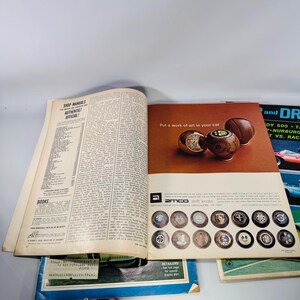 May include: An open magazine showing an advertisement for Amco shift knobs. The ad features three wooden shift knobs and the text "Put a work of art in your car." Various car brand logos are displayed below. The left page is a shop manual.