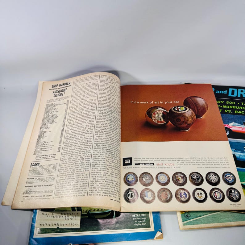 May include: An open magazine showing an advertisement for Amco shift knobs. The ad features three wooden shift knobs and the text "Put a work of art in your car." Various car brand logos are displayed below. The left page is a shop manual.