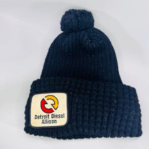 May include: A dark navy blue knit beanie with a pom-pom on top and a wide folded cuff. A rectangular patch is sewn onto the front, featuring a red and yellow circular arrow logo above the black text that reads Detroit Diesel Allison.