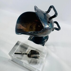 May include: A silver-toned metal coal scuttle shaped sugar bowl with a hinged lid and two handles, accompanied by a small matching metal scoop. The bowl features a pedestal base and a dark, tarnished patina across its curved, polished surface.