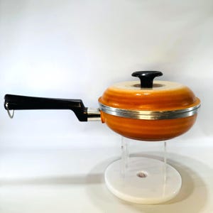 May include: An orange and cream-colored cooking pot with a black handle and lid. The pot has a silver band around the middle and is displayed on a white stand. The handle is angled and has a hole for hanging.