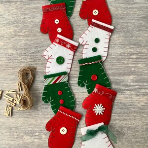 May include: A selection of felt Christmas mitten decorations in red, white, and green. Each mitten is adorned with buttons, ribbon, and stitching. A bundle of twine and small wooden pegs are also present.