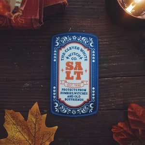 May include: A rectangular patch with a blue border and the text "THE CLEVER-WHITE WITCH CO. SALT EST. 1993 PROTECTS FROM ZOMBIES, WITCHES AND OLD BOYFRIENDS." The patch is surrounded by autumn leaves and a lit candle.