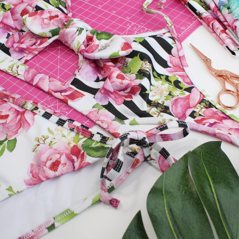 May include: A white swimsuit with black stripes and pink floral print. The swimsuit has a tie at the neck and a tie at the waist. The swimsuit is on a pink cutting mat with a ruler and scissors.