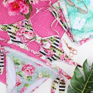May include: A black and white floral print bikini top and matching bottom with a pink floral print. The bikini is laid out on a pink cutting mat with a pair of gold scissors.