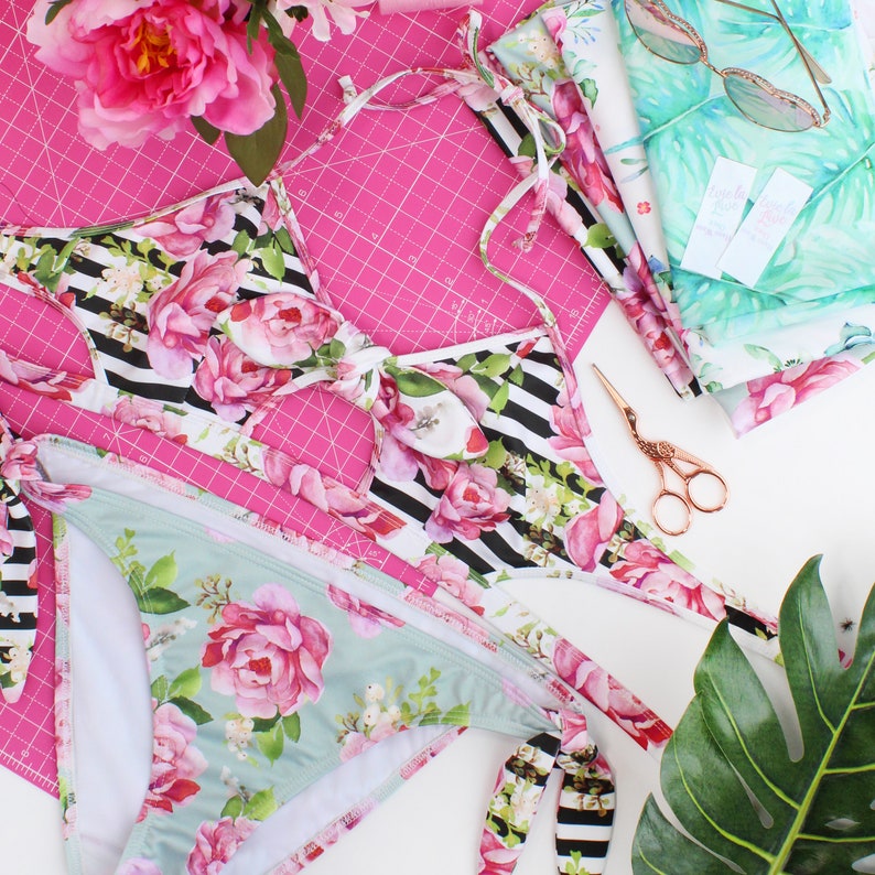 May include: A black and white floral print bikini top and matching bottom with a pink floral print. The bikini is laid out on a pink cutting mat with a pair of gold scissors.