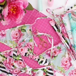 May include: Two pieces of fabric with floral patterns, one with a black and white stripe, and the other with a light blue background. The fabric is laid out on a pink cutting mat with a pair of gold scissors.
