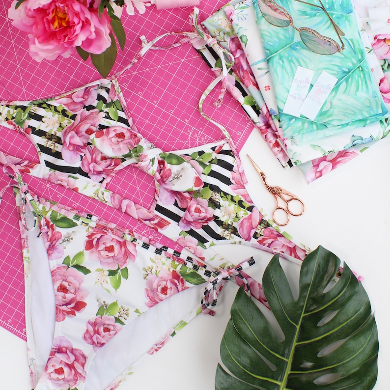 May include: A black and white striped bikini top with pink floral accents and a matching floral print bikini bottom. The bikini is laid out on a pink cutting mat with a green leaf and fabric scraps nearby.