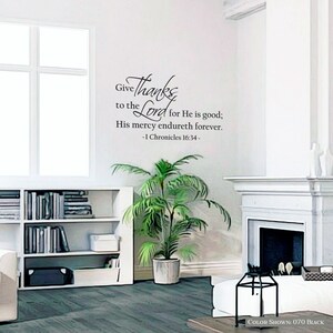 May include: A white wall with a black vinyl decal featuring a bible verse from 1 Chronicles 16:34. The verse reads "Give thanks to the Lord for He is good; His mercy endureth forever." The decal is in a living room setting with a potted plant, a bookshelf, and a fireplace.