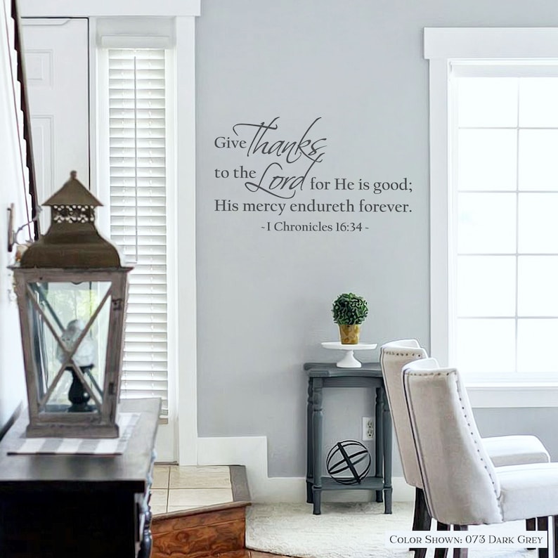 May include: A wall decal with a gray background and a black inscription that reads "Give thanks to the Lord for He is good; His mercy endureth forever. -1 Chronicles 16:34-". The decal is shown in a home setting with a side table, two chairs, and a lantern.