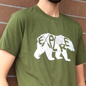 May include: Olive green t-shirt with a white bear graphic. The bear design incorporates the word "EXPLORE" within its outline. The shirt is made of a soft material and has short sleeves.