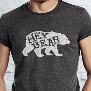 May include: Dark gray t-shirt with rolled sleeves featuring a white graphic of a bear with the words "HEY BEAR" inside the bear's silhouette. The shirt is worn by a person with their hands in their pockets.