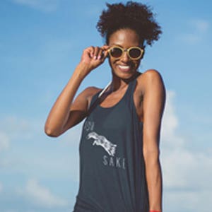May include: A woman wearing a black tank top with the text "For Your Sake" printed on it. She is wearing blue denim shorts and sunglasses. The woman is standing on a pier overlooking the ocean.