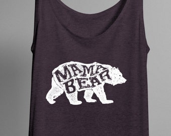 May include: A dark purple tank top with a white graphic of a bear and the words "MAMA BEAR" in a distressed font. The tank top is hanging on a black hanger against a gray background.