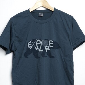 May include: Dark teal t-shirt with the word "EXPLORE" in a stylized font, integrated with a bear silhouette. The design is in a lighter gray color. The shirt is hanging on a wooden hanger against a white background.