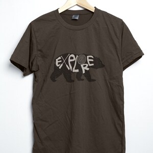 May include: A dark brown t-shirt with the word "EXPLORE" in a white, stylized font, integrated into the silhouette of a bear. The shirt is hanging on a wooden hanger against a white background.