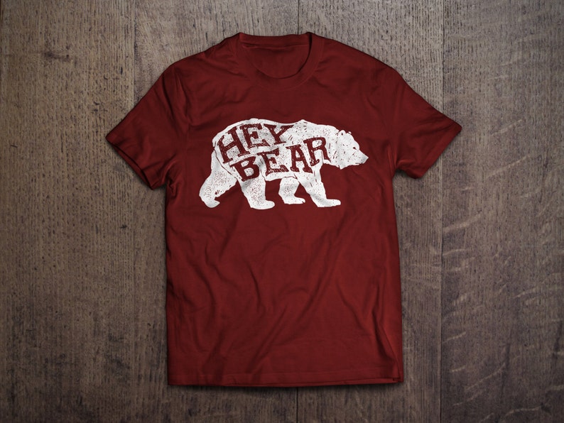 May include: A maroon t-shirt with a distressed white graphic of a bear. The word "HEY" is above "BEAR" inside the bear's silhouette. The shirt is displayed on a wooden surface.