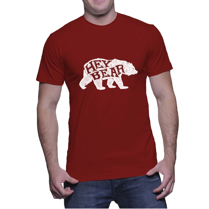 May include: A maroon t-shirt featuring a white bear graphic with the words "HEY BEAR" printed inside the bear's silhouette. The design has a distressed, vintage look. The shirt is short-sleeved and made of a soft material.