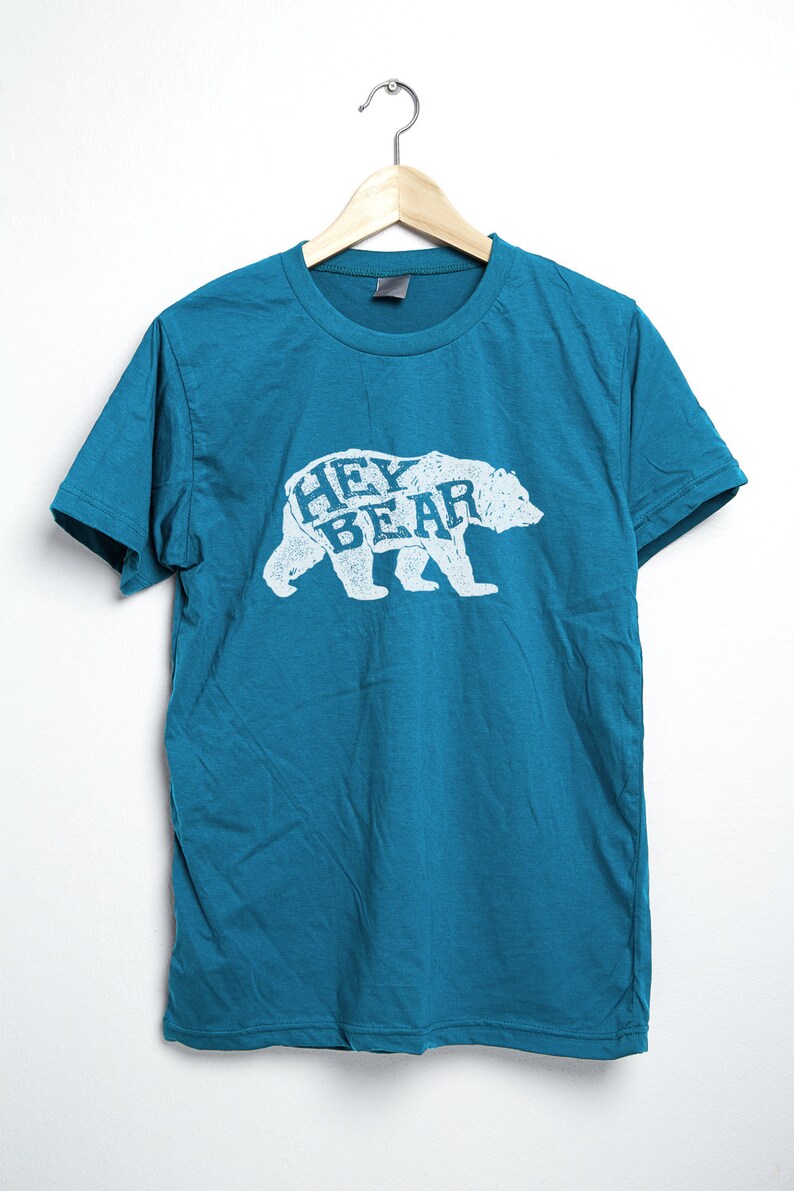 May include: Teal-colored t-shirt with a white graphic of a bear and the words "HEY BEAR" in a distressed font. The shirt is hanging on a wooden hanger against a white background. The t-shirt is made of a soft material.