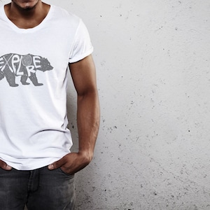 May include: White t-shirt with a gray bear graphic. The word "EXPLORE" is integrated into the bear's silhouette. The shirt has a crew neck and short sleeves.