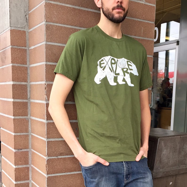 May include: Olive green t-shirt with a white bear graphic. The bear design incorporates the word "EXPLORE" within its outline. The shirt is made of a soft material and has short sleeves.