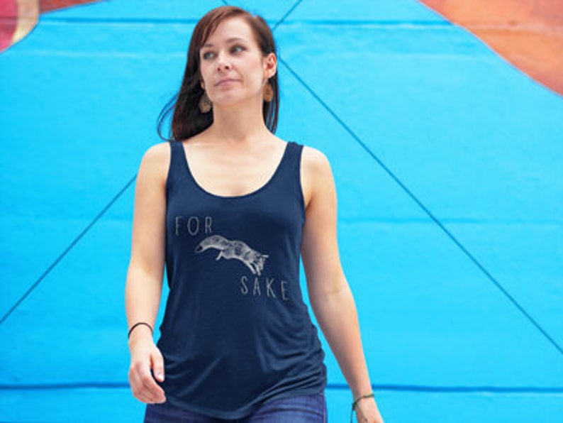 May include: A woman wearing a navy blue tank top with a white graphic of a fox jumping and the text "FOR SAKE".