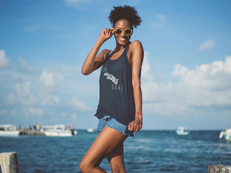 May include: A woman wearing a black tank top with the text "For Your Sake" printed on it. She is wearing blue denim shorts and sunglasses. The woman is standing on a pier overlooking the ocean.