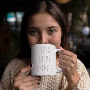 May include: White ceramic mug with black text that reads "Tears of my enemies". A person is holding the mug and drinking from it.