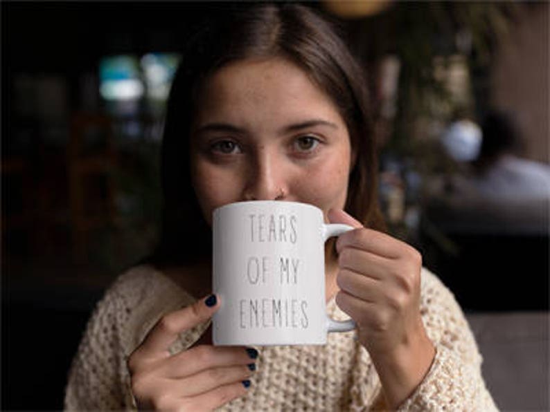 May include: White ceramic mug with black text that reads "Tears of my enemies". A person is holding the mug and drinking from it.