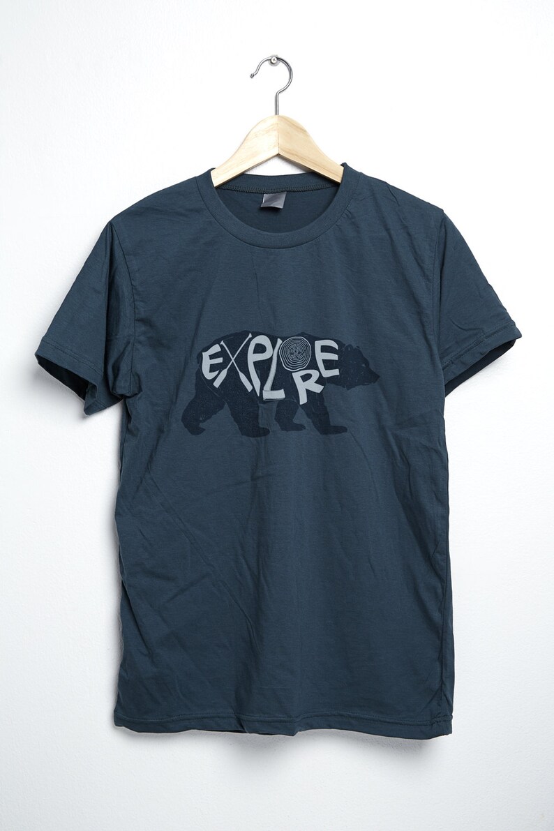 May include: Dark teal t-shirt with the word "EXPLORE" in a stylized font, integrated with a bear silhouette. The design is in a lighter gray color. The shirt is hanging on a wooden hanger against a white background.