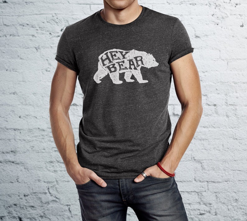 May include: Dark gray t-shirt with rolled sleeves featuring a white graphic of a bear with the words "HEY BEAR" inside the bear's silhouette. The shirt is worn by a person with their hands in their pockets.