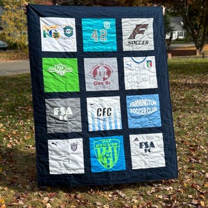 May include: A navy blue quilt with 12 squares, each featuring a different soccer jersey design. The jerseys are in various colors, including white, green, blue, and black. Some jerseys have numbers, logos, and text, such as "FSA FC", "CFC", "PSC Dynamo", and "Farmington Soccer Club".