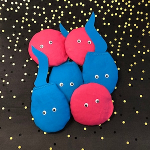 May include: A collection of handmade fabric creatures in shades of red and blue, each with googly eyes. Some have elongated blue appendages. The background is black with gold confetti.