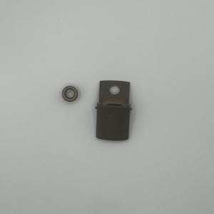 May include: A close-up of two metal hardware pieces. One is a small, round, brass-colored eyelet. The other is a rectangular, brass-colored metal buckle with a small hole in the center.