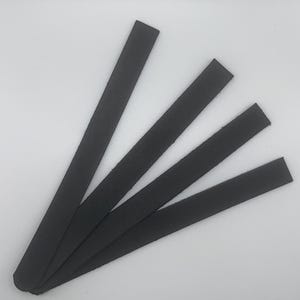 Puede incluir: Cuatro tiras de cuero negro dispuestas diagonalmente sobre un fondo blanco. Cada tira tiene extremos redondeados y una superficie ligeramente texturizada. Las tiras están espaciadas uniformemente. Cada tira mide aproximadamente 1 cm de ancho.