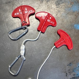 May include: Three red inflation handles with the text "JERK TO INFLATE" and arrows, connected to silver carabiner clips by white cords. The handles are on a dark, textured surface.