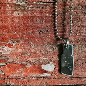 May include: A silver chain necklace with a silver pendant that says "OILER" on a red wooden background.