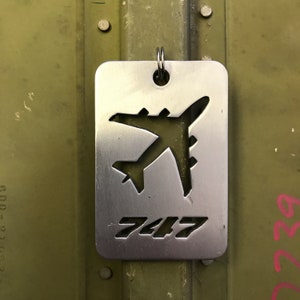 K&ouml;nnte beinhalten: Ein silberner Schl&uuml;sselanh&auml;nger aus Metall mit einem Ausschnitt eines 747-Flugzeugs und der Zahl "747" darunter.