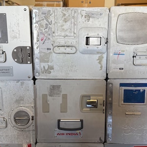 May include: A stack of used, silver-colored airline food service containers. Each container has a handle, latch, and various labels. Some containers have the logos of airlines such as Saudi Arabian Airlines and Air India. The containers are rectangular and appear to be made of metal.