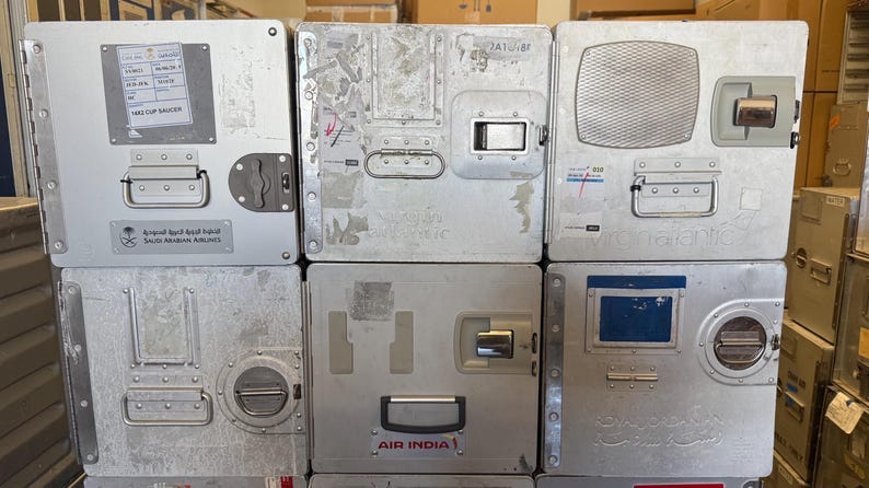 May include: A stack of used, silver-colored airline food service containers. Each container has a handle, latch, and various labels. Some containers have the logos of airlines such as Saudi Arabian Airlines and Air India. The containers are rectangular and appear to be made of metal.