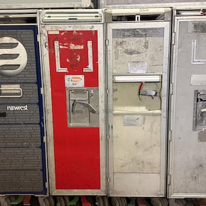 May include: A row of four metal airline service carts. The first cart is dark blue with a white and gray logo and the word "newest". The second cart is red. The other two carts are silver. All carts have wheels.