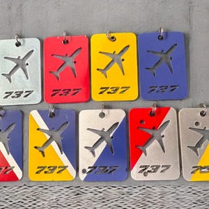 May include: Ten keychains featuring a silver airplane silhouette on a variety of colored backgrounds. The keychains are rectangular and have the number "737" printed on them. The colors include red, blue, yellow, white, and a combination of red, white, and blue.