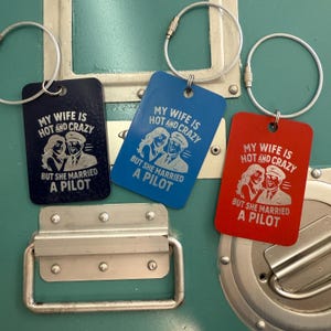 May include: Three rectangular keychains in navy blue, blue, and red. Each keychain has a silver wire loop and features the text "MY WIFE IS HOT AND CRAZY BUT SHE MARRIED A PILOT" with an illustration of a couple.