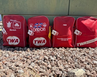 Puede incluir: Cuatro bolsas de viaje TWA vintage rojas con ribetes blancos. Las bolsas tienen diferentes destinos impresos en ellas: Munich, Londres y dos bolsas con solo el logotipo de TWA.