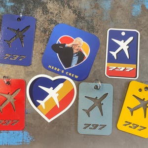 May include: A collection of seven keychains featuring a 737 airplane design. The keychains are made of metal and come in various colours, including red, blue, yellow, and silver. Some keychains feature a heart shape with a 737 airplane design inside. One keychain features a photo of a man in a blue shirt with the text "Herb's Crew".