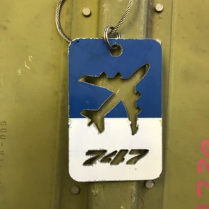 K&ouml;nnte beinhalten: Ein blaues und wei&szlig;es Metallschild mit einer ausgeschnittenen Flugzeugsilhouette und der Nummer "747" auf wei&szlig;em Hintergrund. Das Schild ist an einer silbernen Kette befestigt.