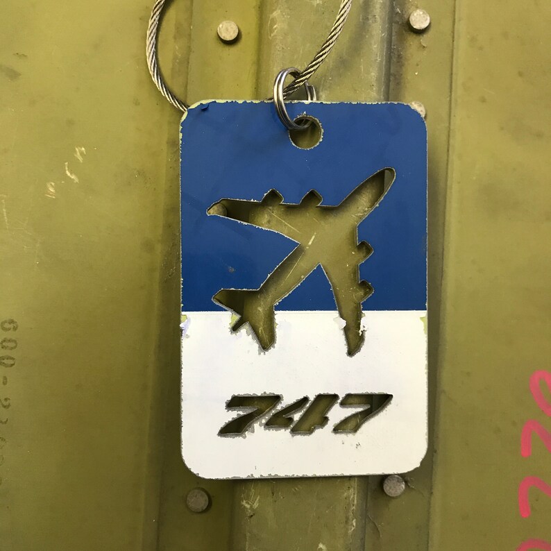 K&ouml;nnte beinhalten: Ein blaues und wei&szlig;es Metallschild mit einer ausgeschnittenen Flugzeugsilhouette und der Nummer "747" auf wei&szlig;em Hintergrund. Das Schild ist an einer silbernen Kette befestigt.