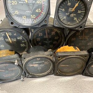 May include: Close-up of eight round analog gauges with black faces and white markings. The gauges measure manifold pressure, hydraulic pressure, and inches of mercury absolute. The gauges are in various sizes and have different labels.