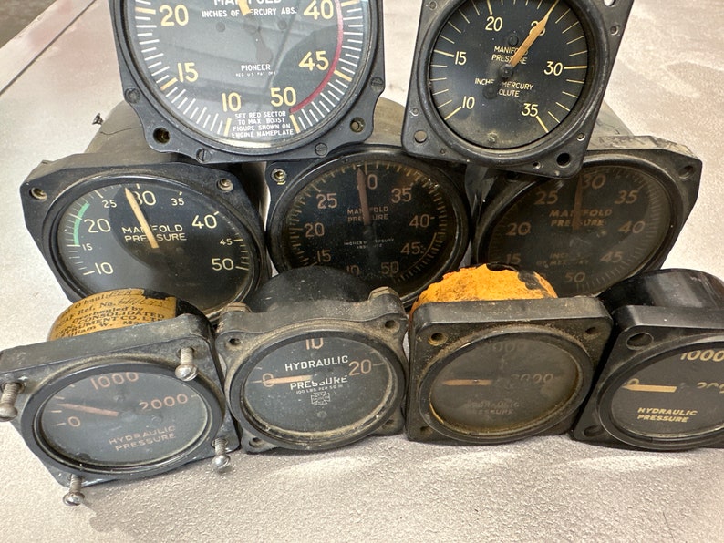 May include: Close-up of eight round analog gauges with black faces and white markings. The gauges measure manifold pressure, hydraulic pressure, and inches of mercury absolute. The gauges are in various sizes and have different labels.